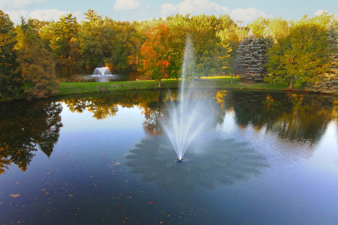An outdoor residential pond with a fountain in the center, surrounded by trees with autumn colors.