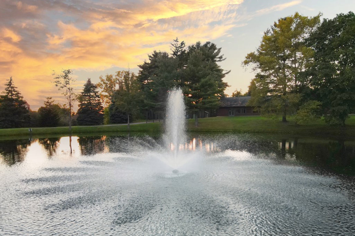 A fountain in operation creating a tall column of water, situated in a pond with a backdrop of trees and a sunset.