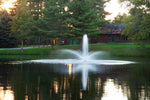 A fountain with multiple spray nozzles in a pond, surrounded by greenery with a house in the background.