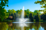 A fountain with a high central jet and wide surrounding spray pattern, situated in a pond with trees in the background.