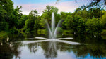 A medium-sized Clover Fountain with a brass center nozzle creating a high center stream and angled spray patterns in a pond, surrounded by trees.