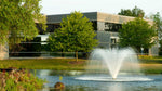 A pond with a water feature created by a display aerator, surrounded by greenery and a building in the background.