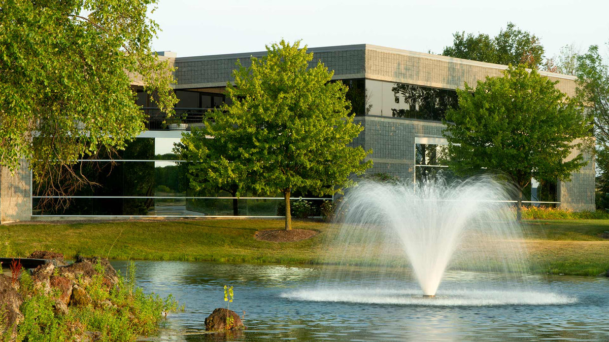 A pond with a water feature created by a display aerator, surrounded by greenery and a building in the background.