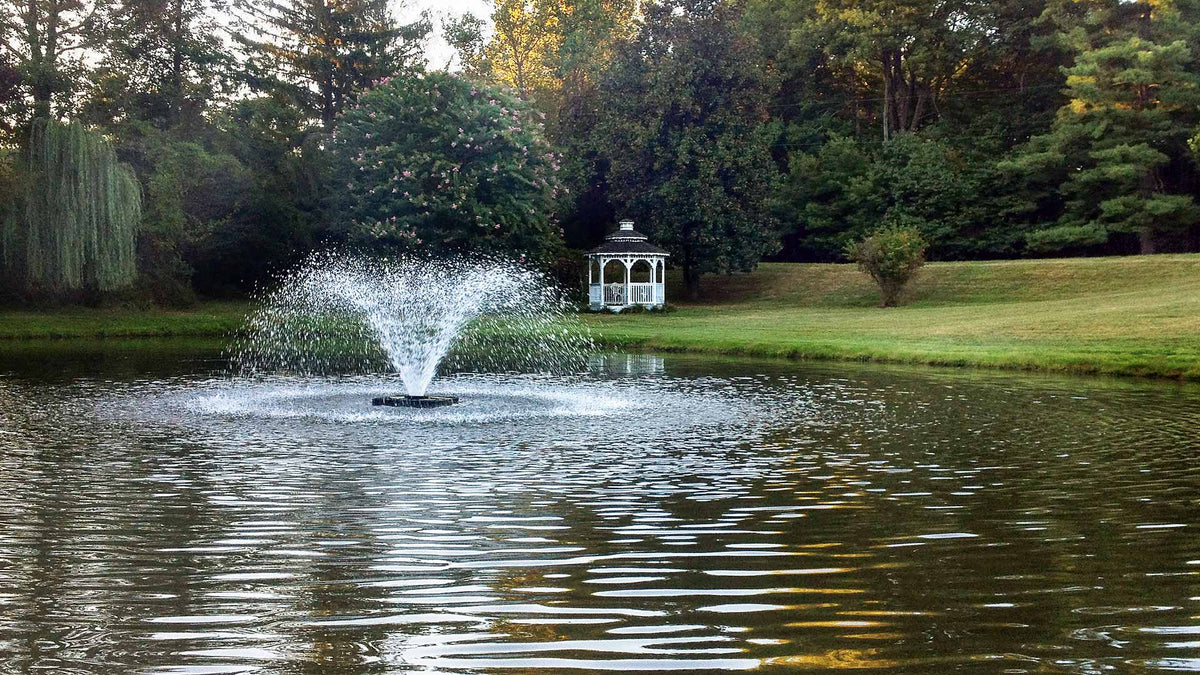 Fountain in a pond with a gazebo in the background, surrounded by trees and greenery.